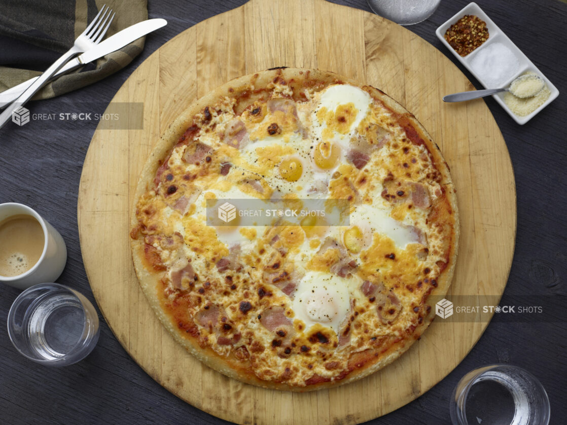 Whole breakfast pizza with bacon and eggs on a round wood pizza peel, grey wood background, overhead