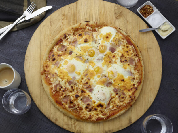 Whole breakfast pizza with bacon and eggs on a round wood pizza peel, grey wood background, overhead