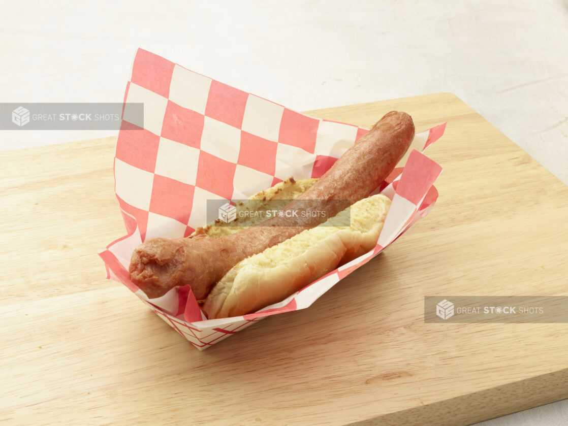 Plain hot dog on a bun in a red and white cardboard takeout container lined with red and white checkered paper liner