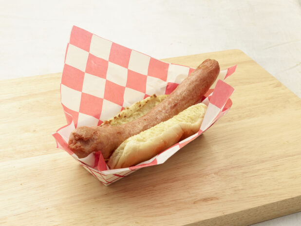 Plain hot dog on a bun in a red and white cardboard takeout container lined with red and white checkered paper liner