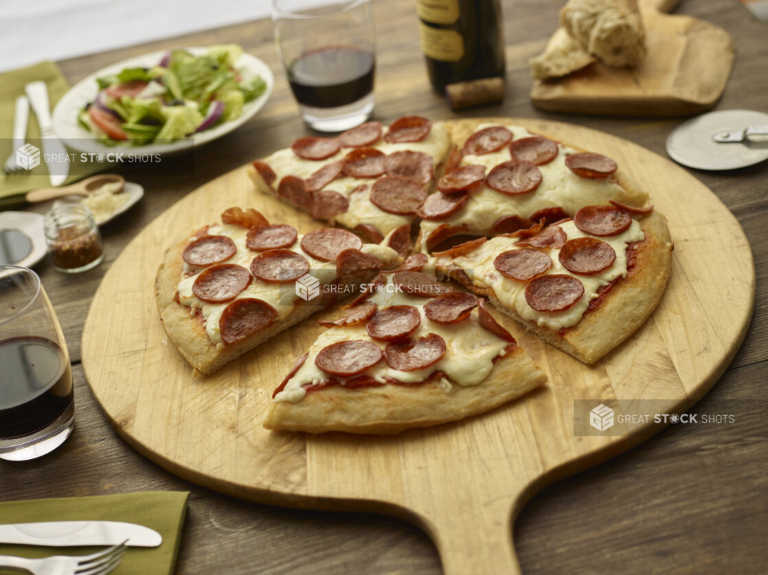 Sliced pepperoni pizza on a round wood pizza peel with pizza cutter, wooden tabletop, salad and glasses of wine surrounding