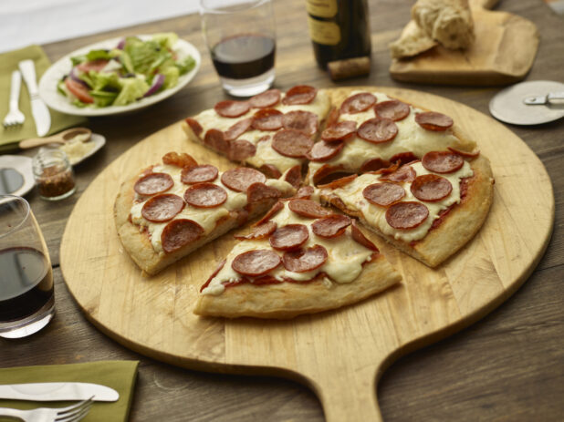 Sliced pepperoni pizza on a round wood pizza peel with pizza cutter, wooden tabletop, salad and glasses of wine surrounding
