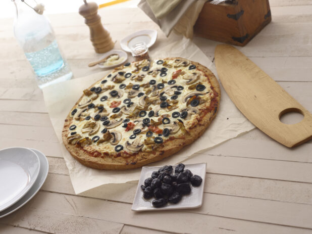 Whole 3-topping pizza with mushrooms, black olives, and hot peppers on parchment with a small bowl of dried black olives, side plates, and condiments surrounding