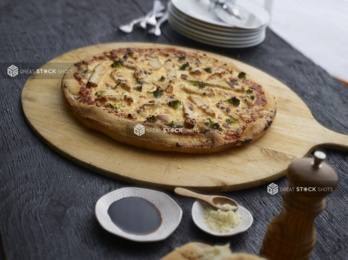 Whole chicken, broccoli, and cheese pizza on a wood pizza peel with cutter, condiments and tableware surrounding, black background