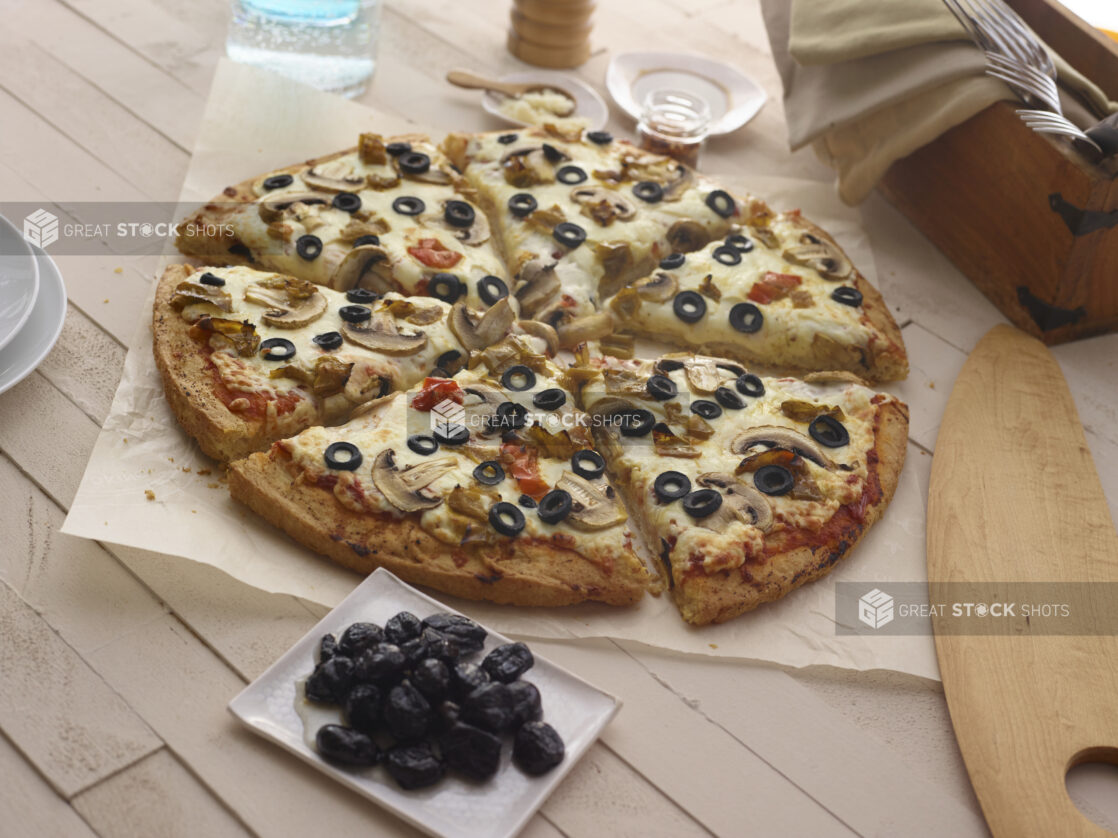 Whole 3-topping pizza with mushrooms, black olives, and hot peppers on parchment with a small bowl of dried black olives,