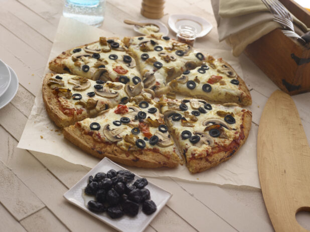 Whole 3-topping pizza with mushrooms, black olives, and hot peppers on parchment with a small bowl of dried black olives,