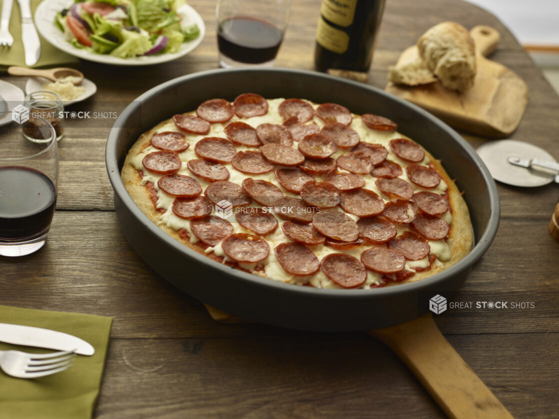 Whole unsliced pepperoni pan pizza on a wood peel on a wood tabletop with torn bread, red wine, green salad, and condiments surrounding