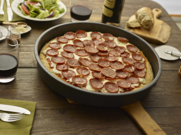 Whole unsliced pepperoni pan pizza on a wood peel on a wood tabletop with torn bread, red wine, green salad, and condiments surrounding