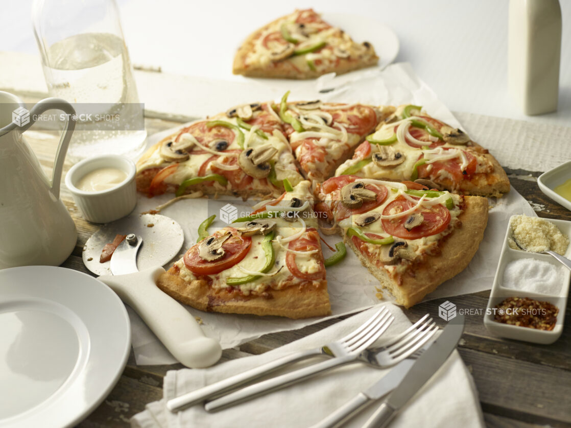 Sliced veggie deluxe pizza with tomatoes, onions, mushrooms, and green peppers on white parchment with utensils and condiments surrounding