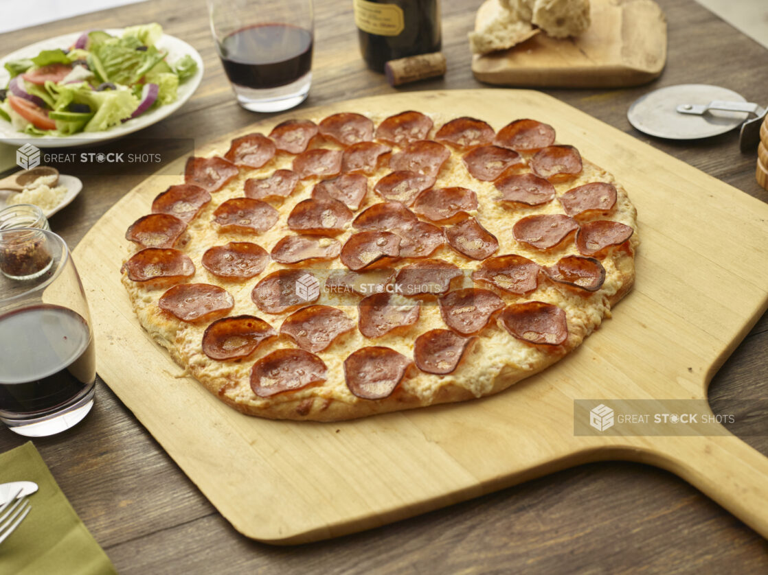 Whole pepperoni pizza on a pizza peel accompanied by side salad, red wine, pizza cutter, chili flakes