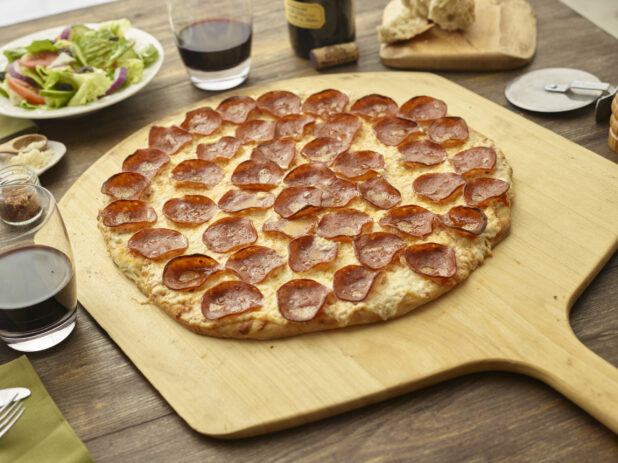 Whole pepperoni pizza on a pizza peel accompanied by side salad, red wine, pizza cutter, chili flakes