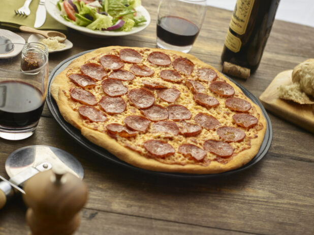 Whole pepperoni pizza on a round wood pizza peel, wooden tabletop, salad and glasses and bottle of wine surrounding