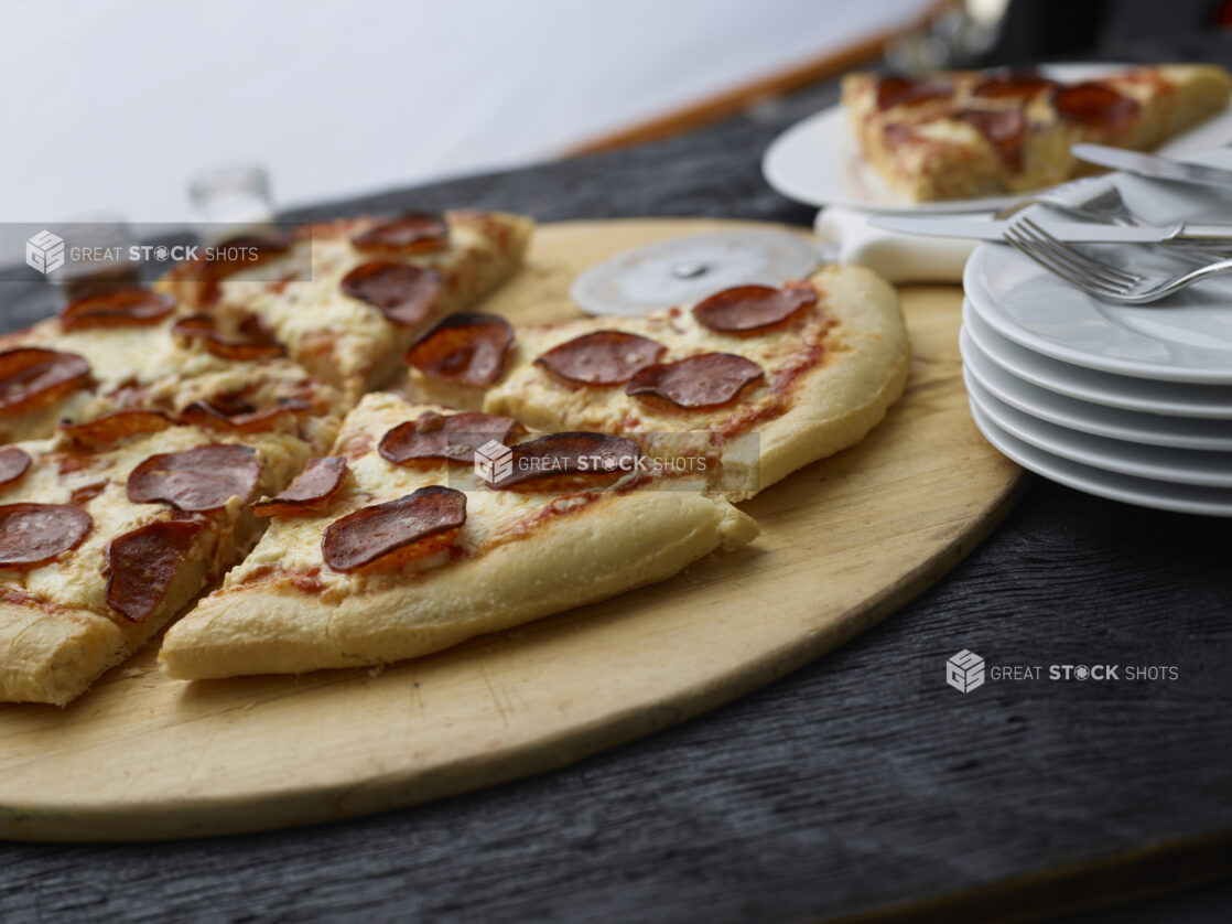 Sliced pepperoni pizza on a wood pizza peel, side plate alongside, black tabletop