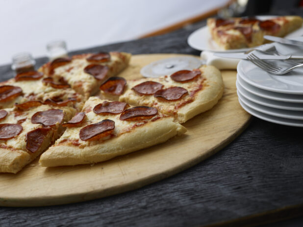 Sliced pepperoni pizza on a wood pizza peel, side plate alongside, black tabletop