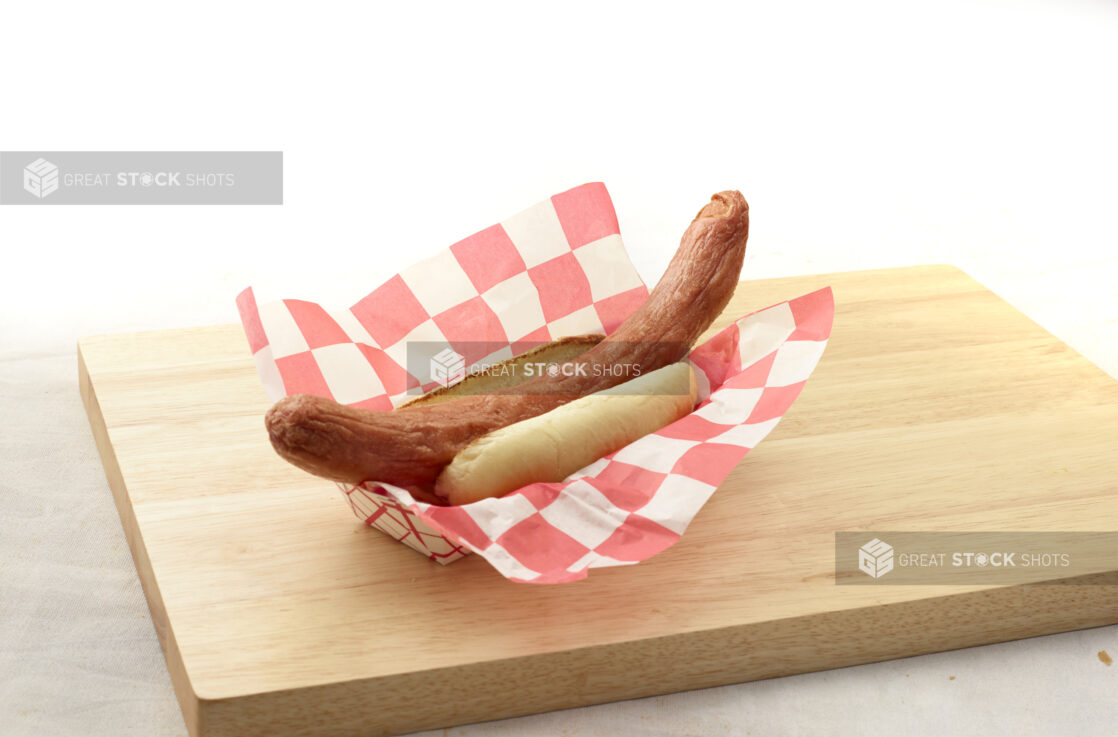 Long hot dog on a bun in a red and white lined cardboard takeout container on a wood board, white background