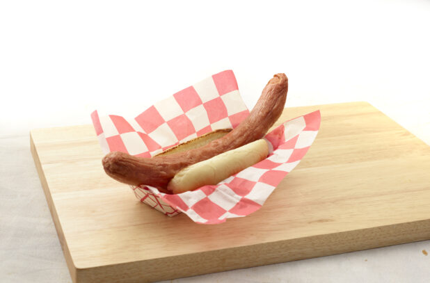 Long hot dog on a bun in a red and white lined cardboard takeout container on a wood board, white background