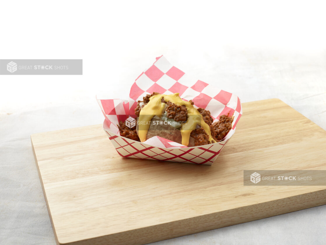 Baked potato with chilli and cheese sauce in a red and white lined cardboard takeout container on a wood board, white background