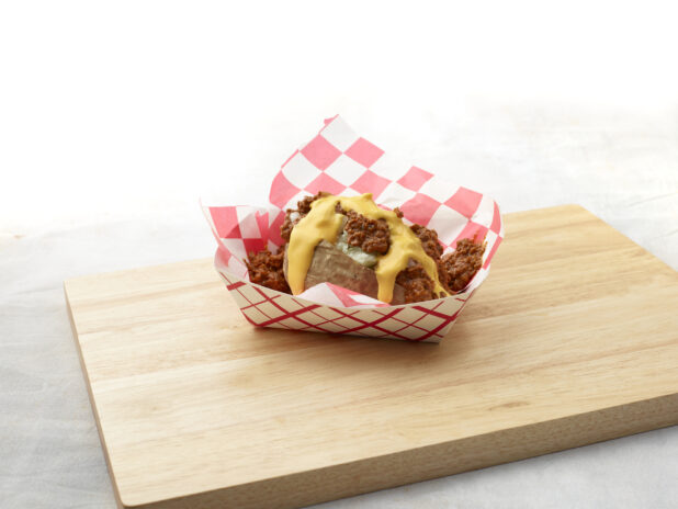 Baked potato with chilli and cheese sauce in a red and white lined cardboard takeout container on a wood board, white background