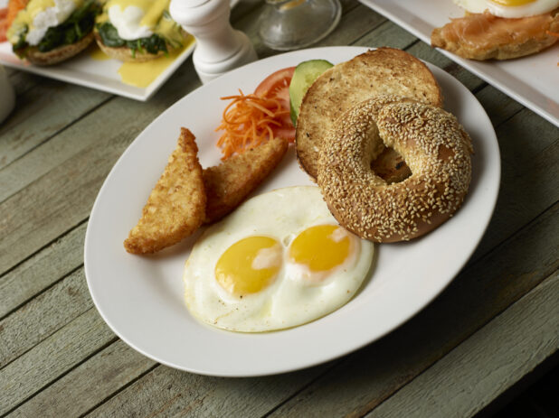Two eggs sunny side up on a white plate with a toasted sesame bagel, hash browns, and veggie garnish, green wood background