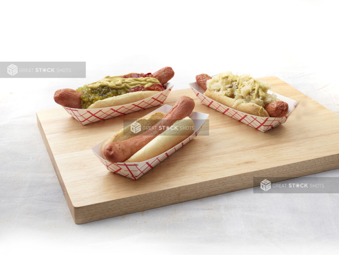 Three hot dogs with various toppings in red and white takeout containers on a wooden board, white background