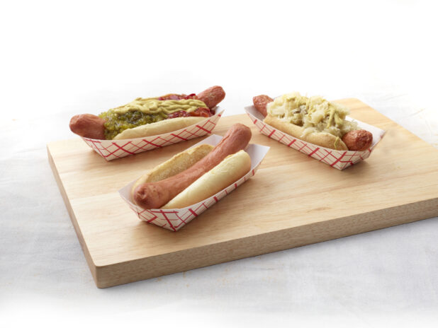 Three hot dogs with various toppings in red and white takeout containers on a wooden board, white background