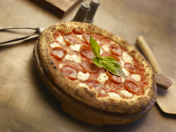 Whole personal-size Neapolitan-style pepperoni pizza with fresh basil garnish on a round wooden block, wood background