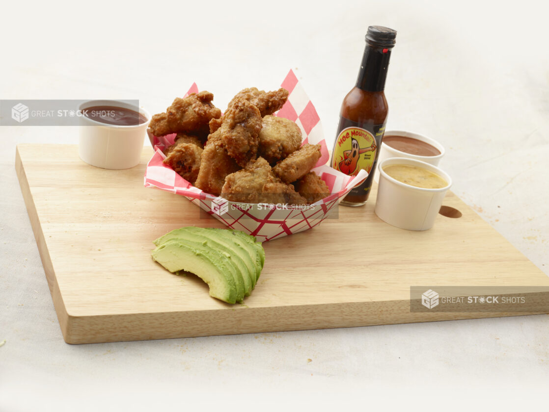 Breaded fried chicken wings in a red and white lined takeout container, dips and avocado slices alongside, on a wood board, white background