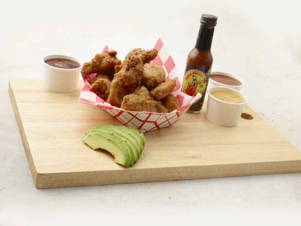 Breaded fried chicken wings in a red and white lined takeout container, dips and avocado slices alongside, on a wood board, white background