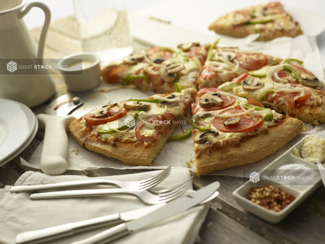 Sliced veggie deluxe pizza with tomatoes, onions, mushrooms, and green peppers on white parchment with utensils and condiments surrounding