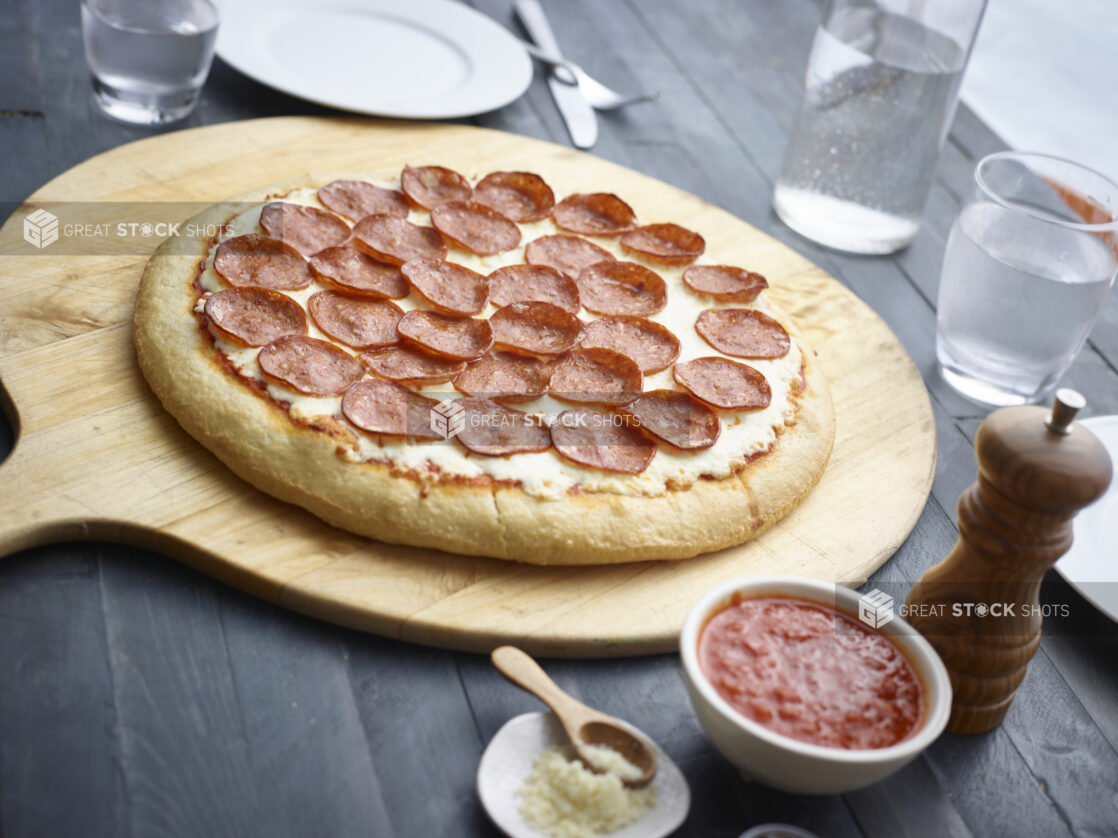 Whole pepperoni pizza on a wood pizza peel, condiments and water glass alongside, all on a black wood tabletop