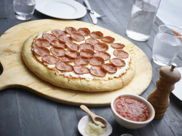 Whole pepperoni pizza on a wood pizza peel, condiments and water glass alongside, all on a black wood tabletop