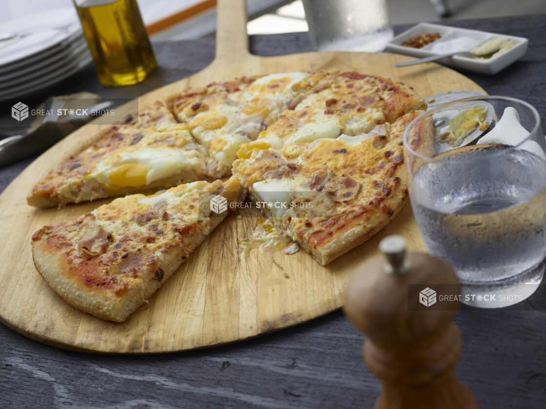 Whole breakfast pizza with bacon and eggs on a round wood pizza peel, slice removed, grey wood background, tilted angle