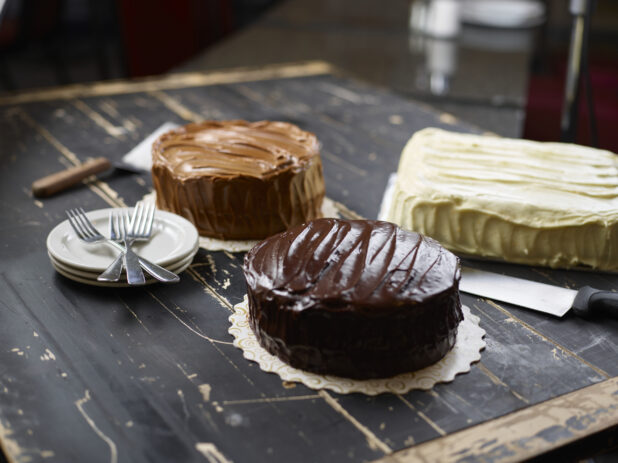 A variety of iced cakes - caramel, chocolate, and vanilla - with side plates and forks on a black wood tabletop