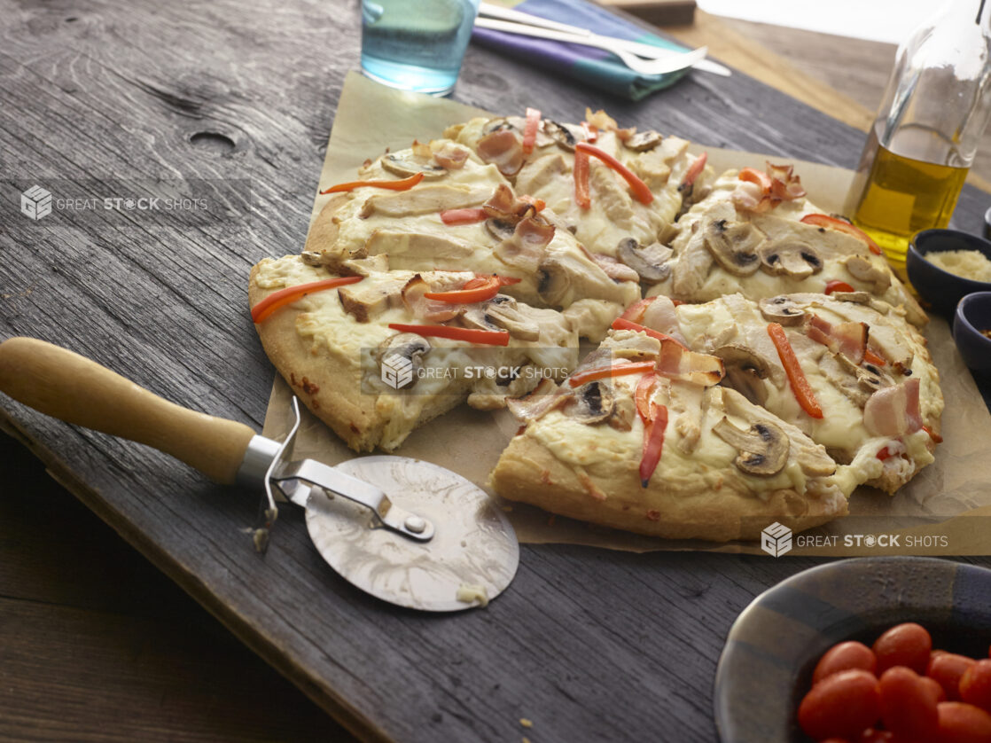 Sliced bacon, mushroom, and red pepper pizza with a cutter on parchment paper, table items surrounding, wood tabletop