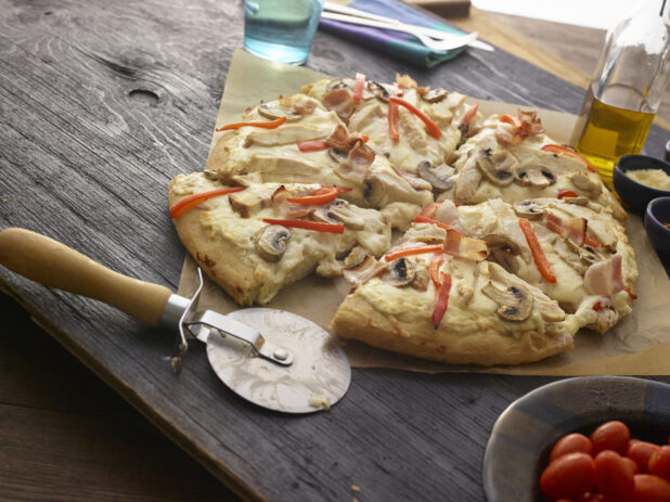 Sliced bacon, mushroom, and red pepper pizza with a cutter on parchment paper, table items surrounding, wood tabletop