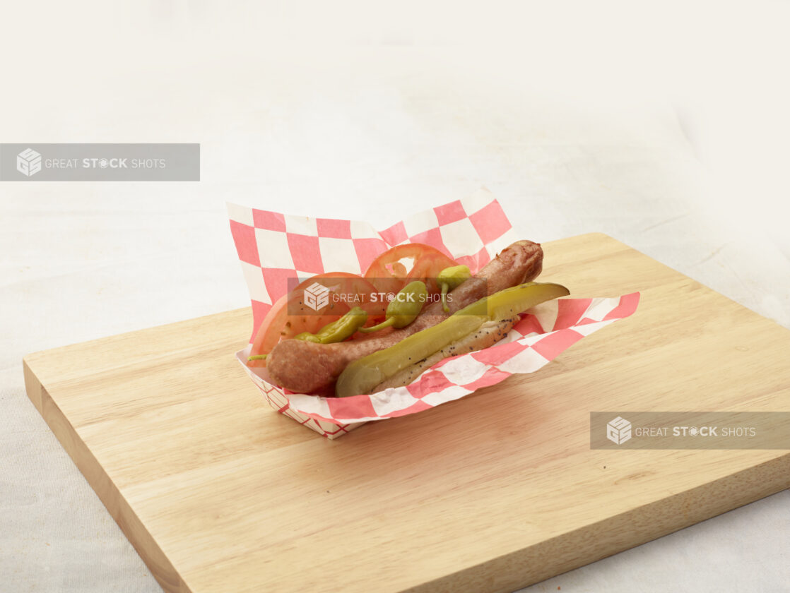 Ballpark-style frank on a poppyseed bun with dill pickle spear, sliced tomatoes, and pickled peppers in a cardboard takeout container on a wood board, white background