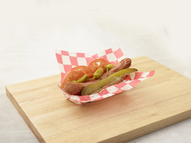 Ballpark-style frank on a poppyseed bun with dill pickle spear, sliced tomatoes, and pickled peppers in a cardboard takeout container on a wood board, white background