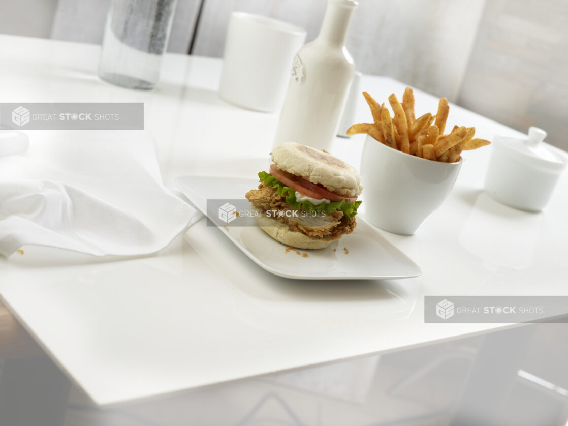 Crispy chicken sandwich on an English muffin with tomato, lettuce, and mayo, french fries in a white ceramic cup on the side, white background