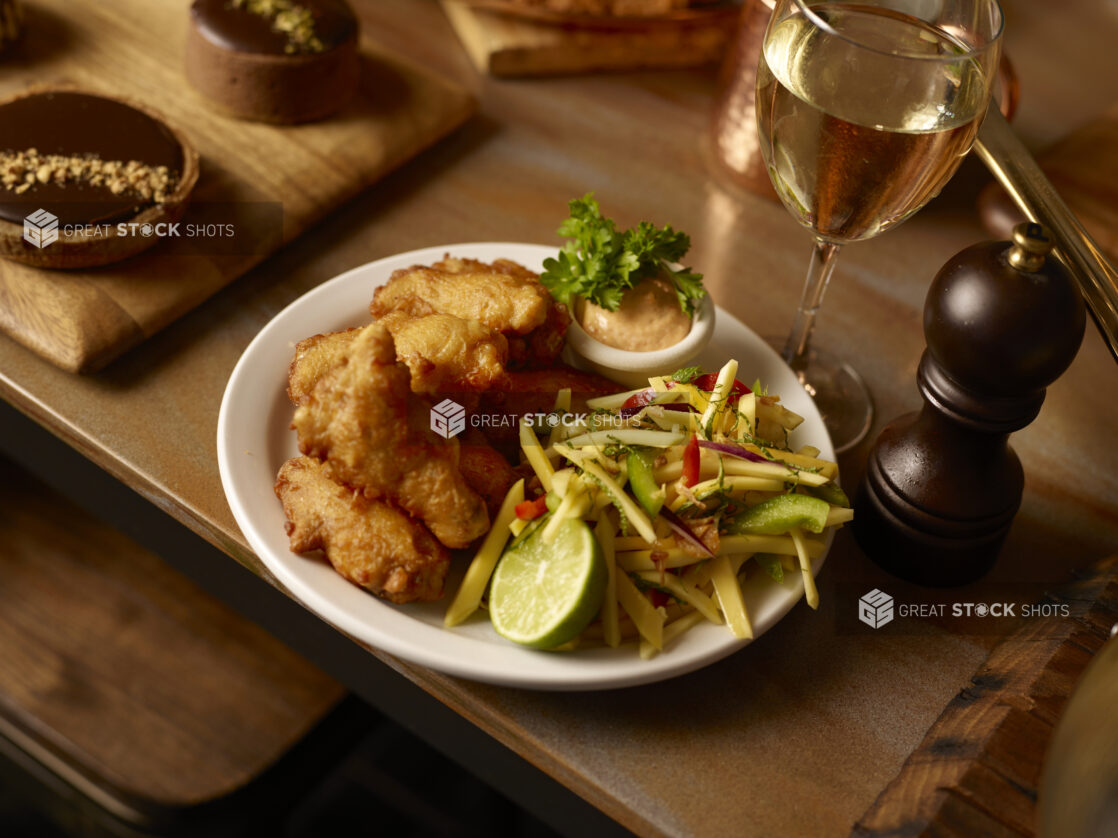 Nuggets of battered fried fish on a white plate with shredded Thai-style mango salad, pepper mill, glass of white wine, and chocolate tart surrounding on wood tabletop