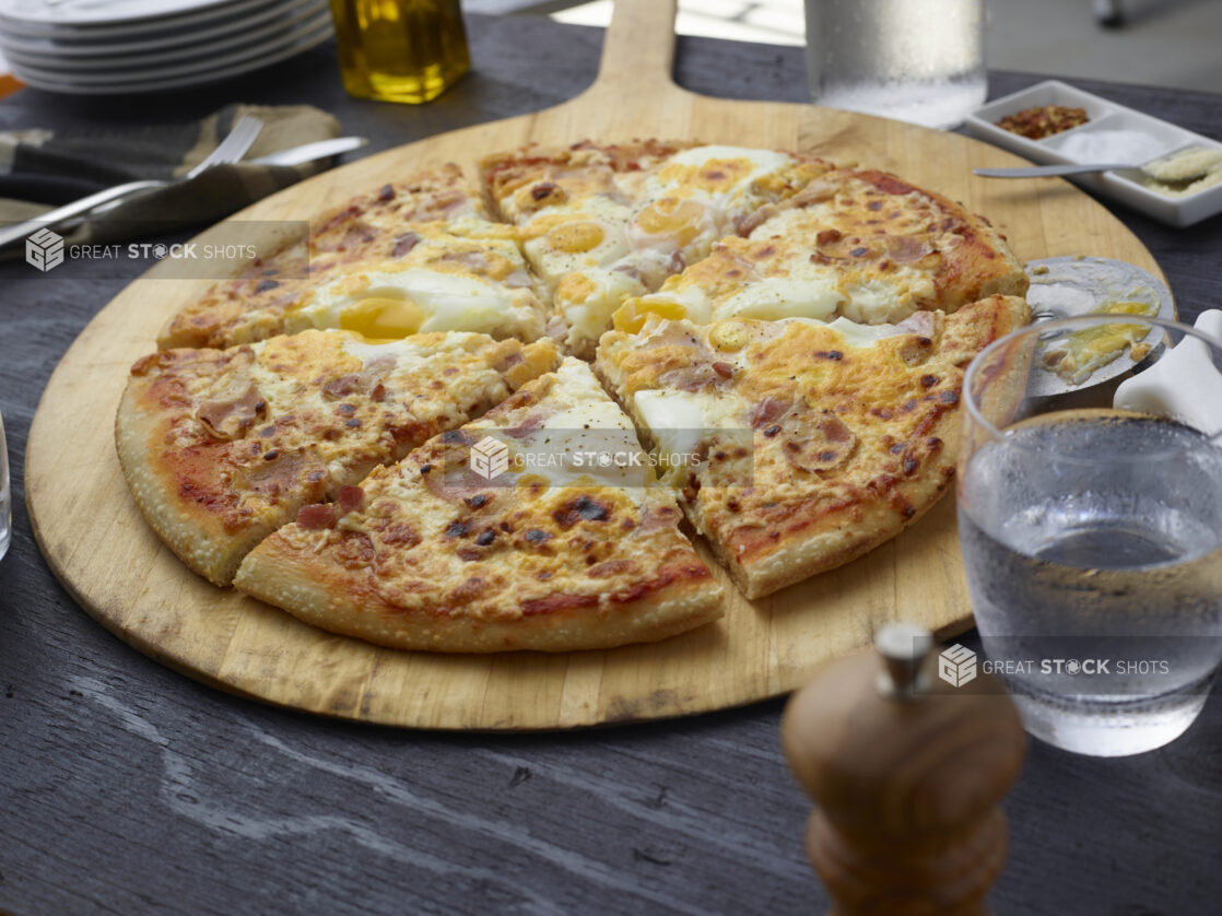 Sliced breakfast pizza with bacon and eggs on a round wood pizza peel, grey wood background,