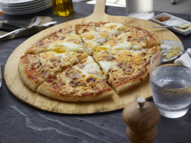 Sliced breakfast pizza with bacon and eggs on a round wood pizza peel, grey wood background,