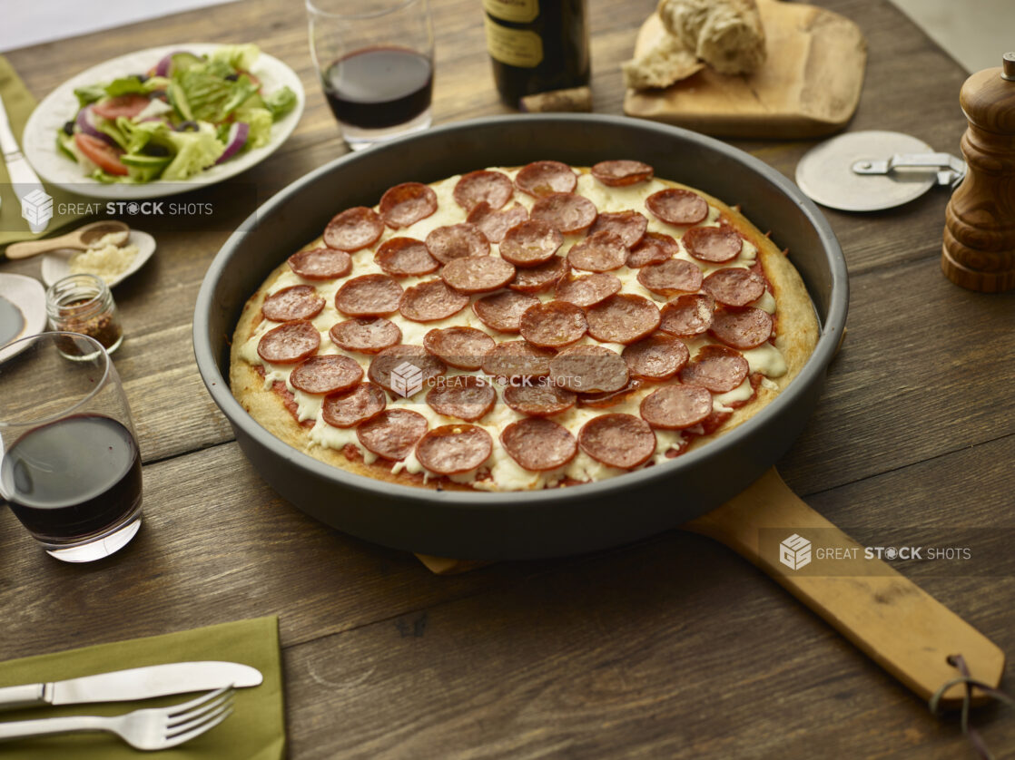 Whole unsliced pepperoni pan pizza on a wood peel on a wood tabletop with torn bread, red wine, green salad, and condiments