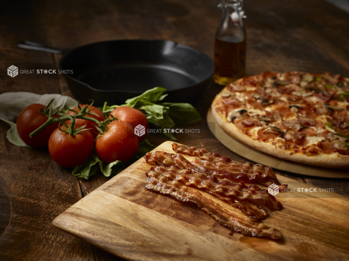 Strips of crispy bacon on a wood board, vine tomatoes, skillet, and whole meat and mushroom pizza in background, wood background