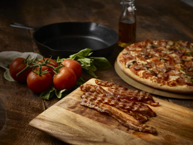 Strips of crispy bacon on a wood board, vine tomatoes, skillet, and whole meat and mushroom pizza in background, wood background