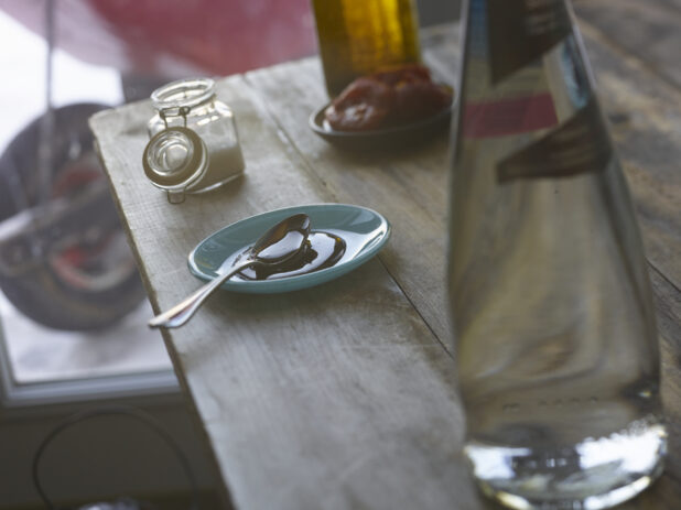 A Teaspoon in a Teal Ceramic Dish with Olive Oil and a Jar of Salt on a Wooden Table in a Kitchen Setting