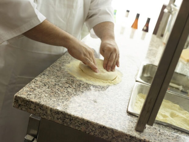 Man kneading pizza dough in a white apron in a pizza kitchen on marble