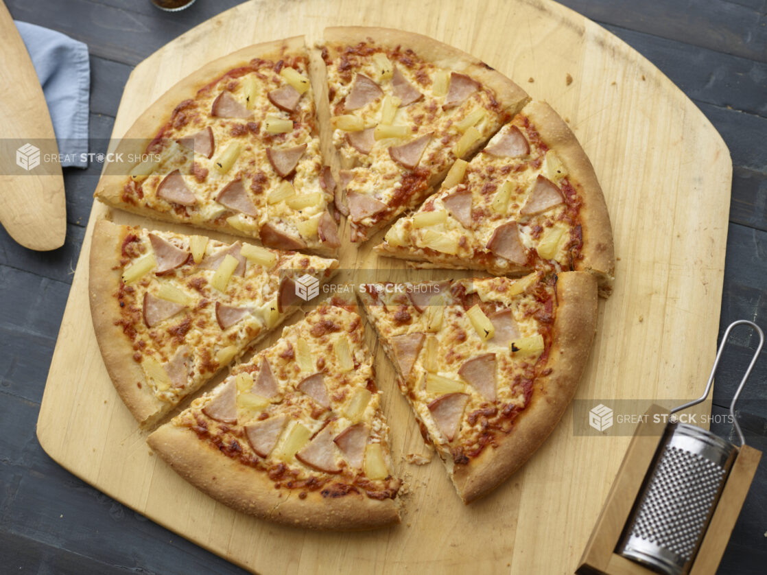 Whole sliced Hawaiian pizza on a wood pizza peel, parmesan grater alongside, black wood background