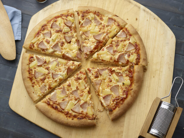 Whole sliced Hawaiian pizza on a wood pizza peel, parmesan grater alongside, black wood background