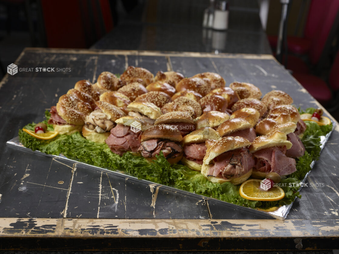 Assorted miniature deli meat sandwiches on sesame seed rolls on a lettuce-lined serving tray, distressed black painted wood background