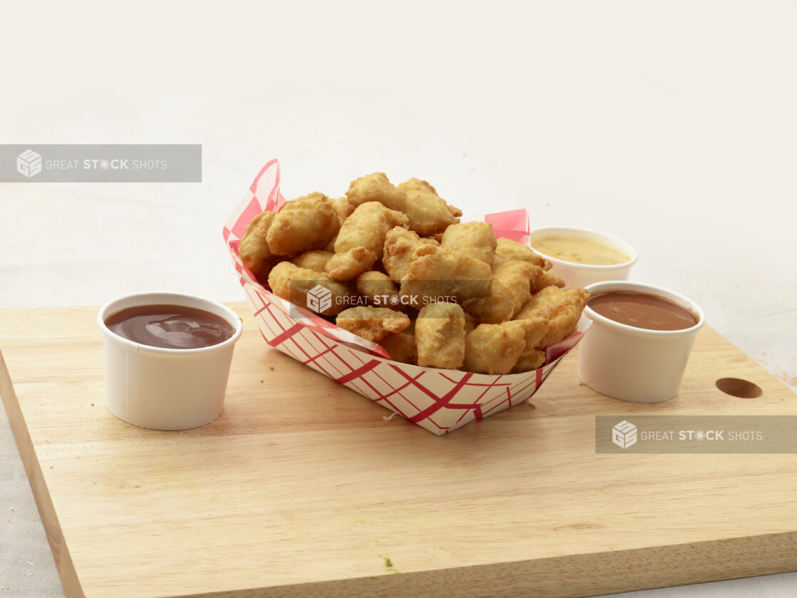 Battered chicken nuggets in a red and white cardboard takeout container, dipping sauces alongside, on a wood board, white background